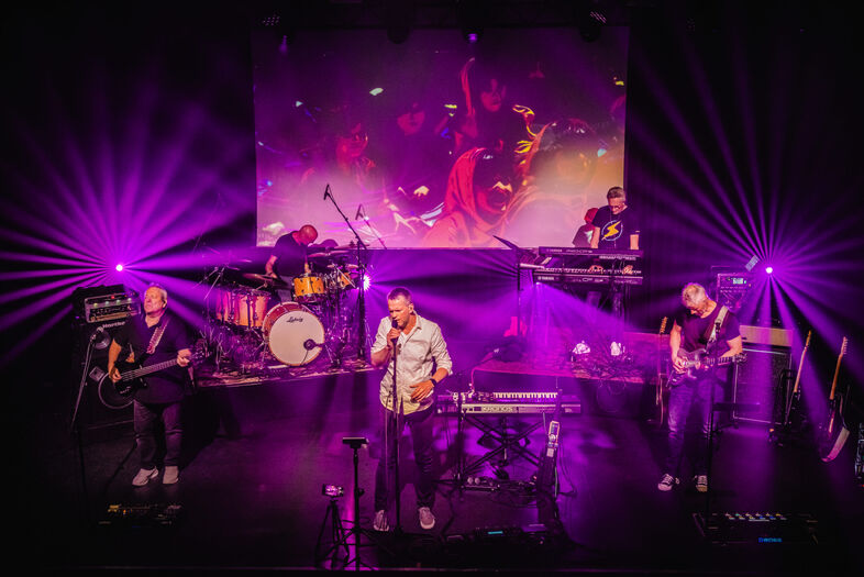 Een band speelt live op het podium met kleurrijke lichten en een visuele achtergrondprojectie.
