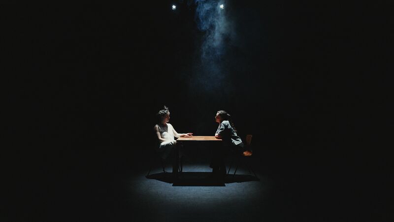 Twee personen zitten tegenover elkaar aan een tafel in een donker theater, verlicht door een enkele spot.