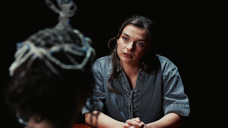 Een vrouw met lange, donkere haren zit aan een tafel en kijkt aandachtig naar iemand buiten beeld.