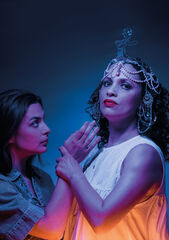 Twee vrouwen in dramatische pose, met kleurrijke verlichting; één in toga en hoofdtooi, de ander in casual kleding.