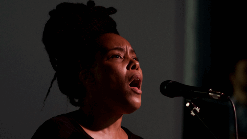 Elaine Mitchener zingt in een microfoon met een intense expressie tijdens een optreden.