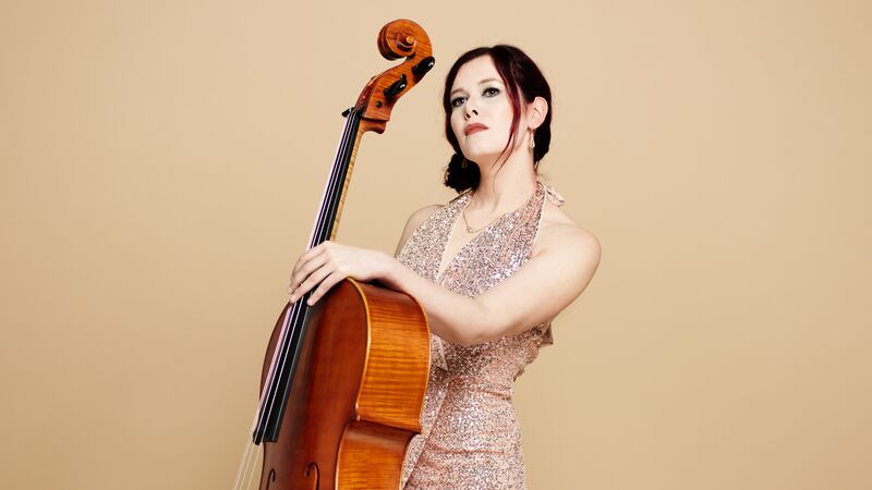 Vrouw in een glinsterende jurk houdt een cello vast en kijkt naar de camera tegen een neutrale achtergrond.