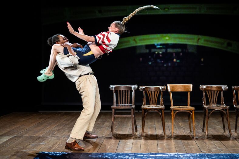 Een man tilt een kind in de lucht op een podium met stoelen op de achtergrond, terwijl ze beiden lachend zijn.