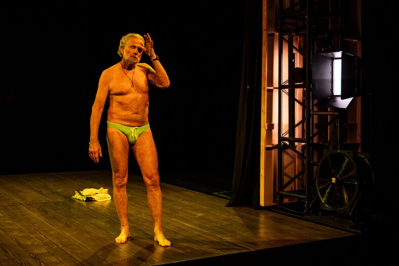 Een naakte performer staat op een podium onder een felle lichten, met een reflectie van een andere kledingstuk op de grond.