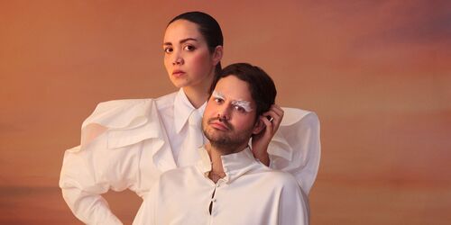 Man en vrouw in witte, sculpturale kleding poseren statig voor een zachtroze avondlucht, waarbij de vrouw achter de man staat met één hand op haar heup.