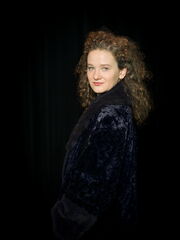 Portret van een vrouw met krullend haar, gekleed in een zwarte jas, tegen een donkere achtergrond.