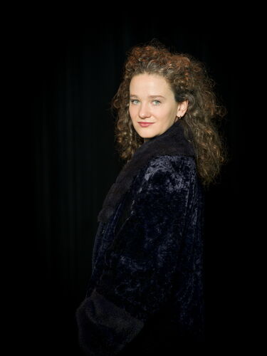 Portret van een vrouw met krullend haar, gekleed in een zwarte jas, tegen een donkere achtergrond.