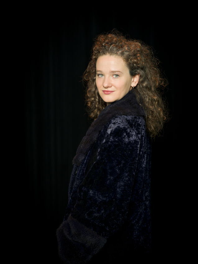 Portret van een vrouw met krullend haar, gekleed in een zwarte jas, tegen een donkere achtergrond.