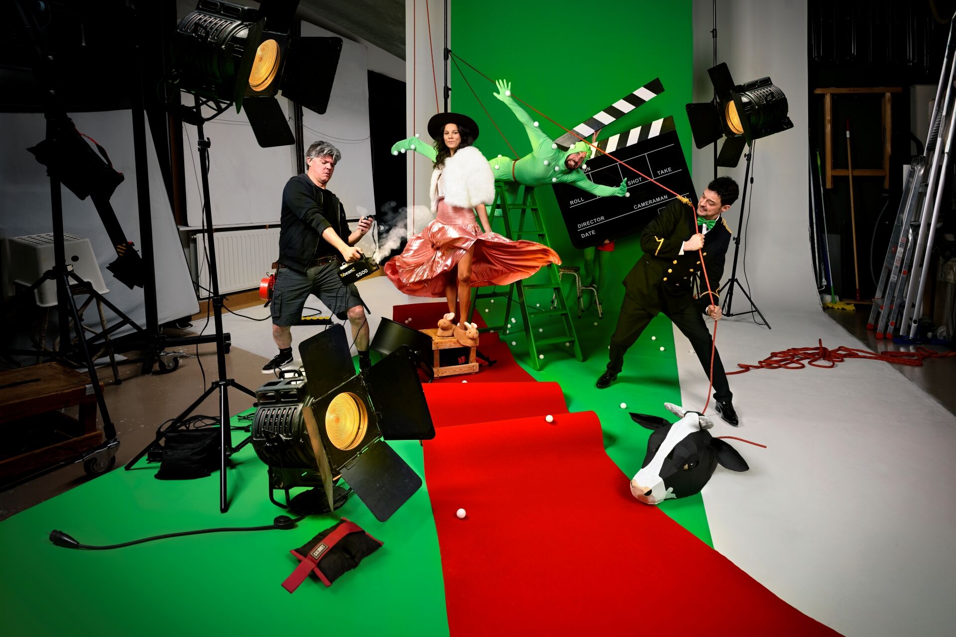 Filmset met groene achtergrond, rode loper, meerdere personen, camera‑lampen en rekwisieten.