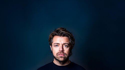 Frontaal portret van een persoon met licht golvend haar tegen een donkerblauwe achtergrond.