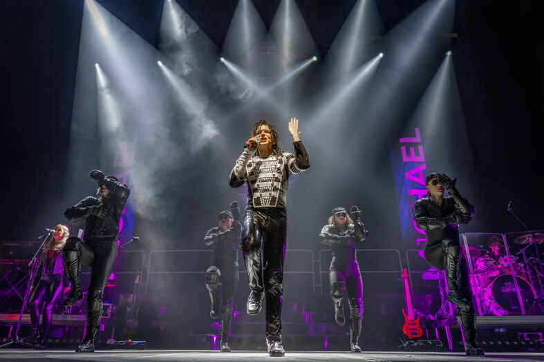 Een zanger performing op het podium met dansers achter hem, verlicht door felle lichten.