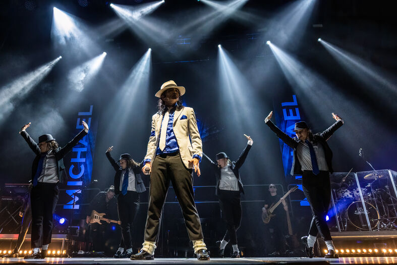 Een zanger in een beige pak staat in het middelpunt van het podium, omringd door dansers in zwart-witte outfits.