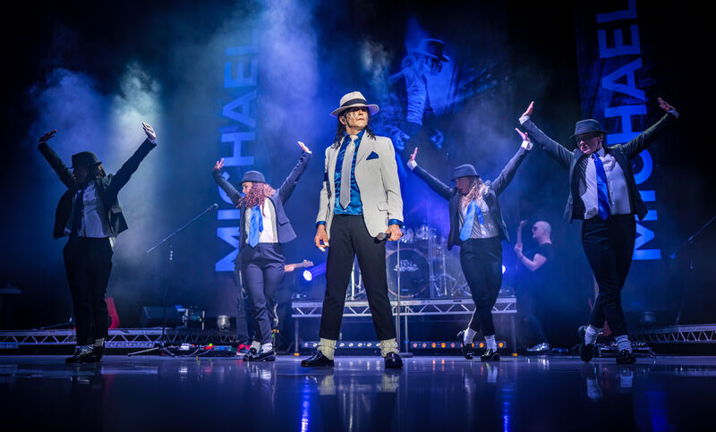Optreden van een zanger in een wit jasje met dansers op een podium, omringd door kleurrijke lichten en grote blauwe tekst.