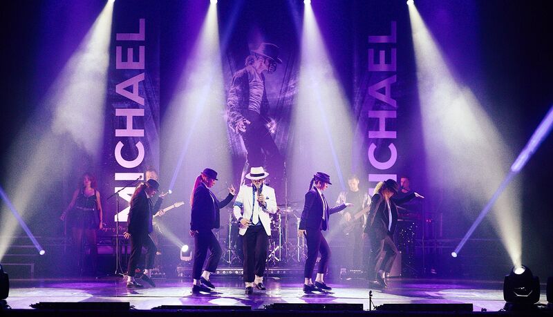 Een live-optreden met dansers in zwarte outfits en een zanger in het midden, verlicht door paarse en witte lampen.