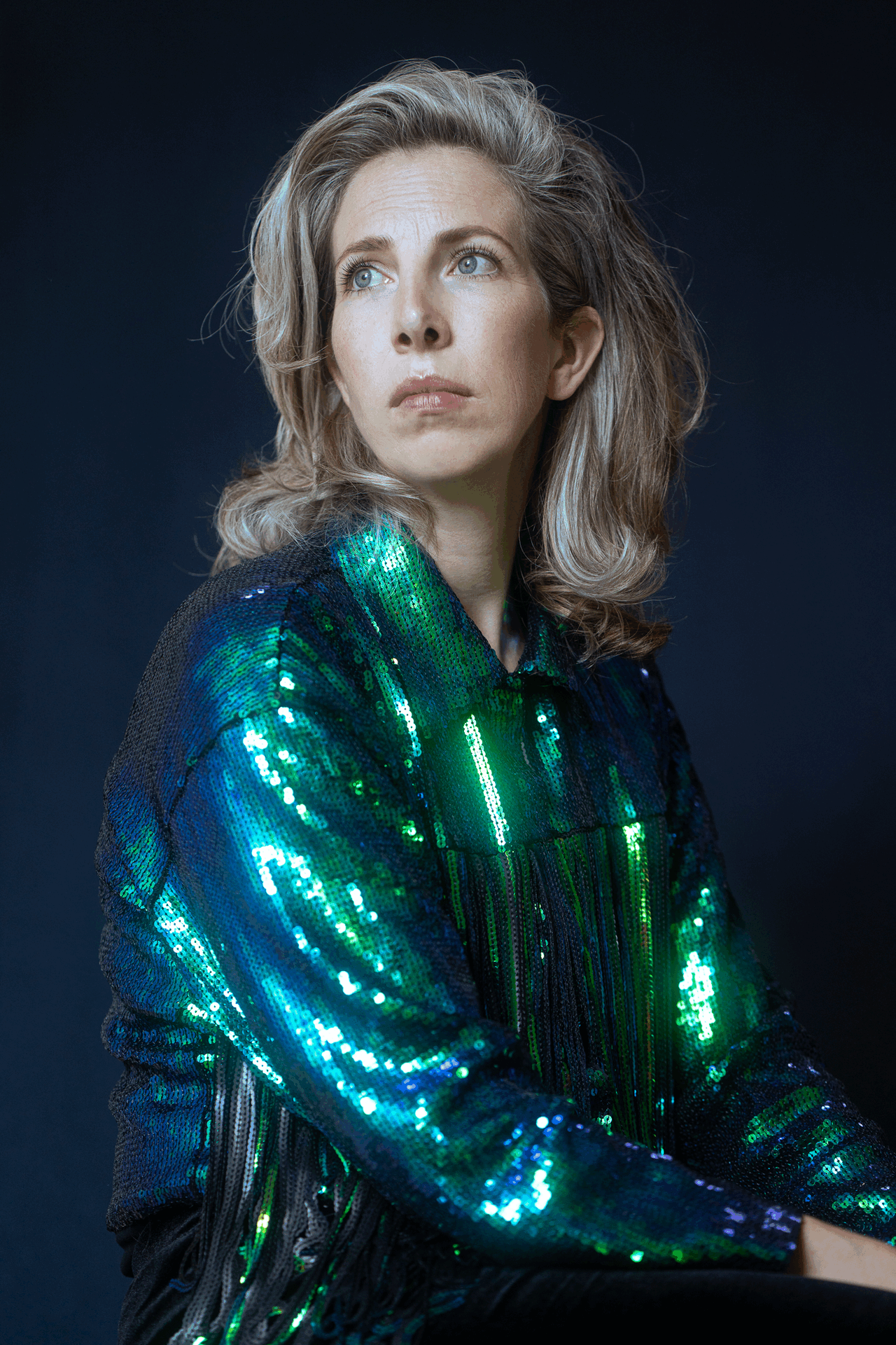 Vrouw met een glinsterende blauw-groene blouse, zittend en kijkend naar de zijkant tegen een donkere achtergrond.