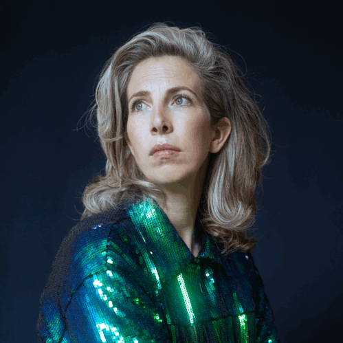 Vrouw met een glinsterende blauw-groene blouse, zittend en kijkend naar de zijkant tegen een donkere achtergrond.