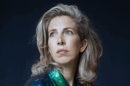 Vrouw met een glinsterende blauw-groene blouse, zittend en kijkend naar de zijkant tegen een donkere achtergrond.