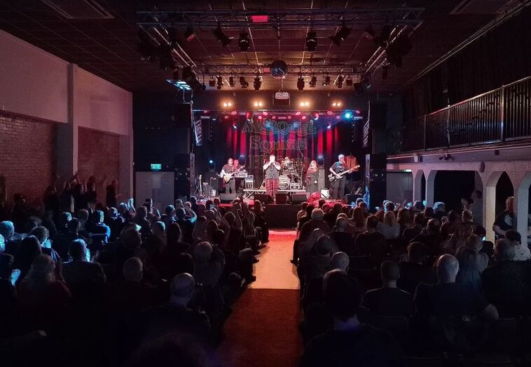 Een concert met een band op het podium, omringd door een publiek in een sfeervolle zaal met dim licht.