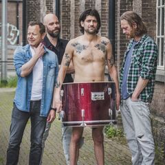 Vier personen staan in een steeg; één shirtloos met grote trom voor lichaam.