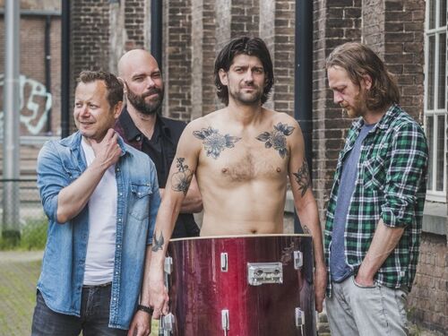 Vier personen staan in een steeg; één shirtloos met grote trom voor lichaam.