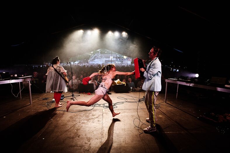 Live optreden van een muzikale act met een danseres en twee performers op een podium voor een enthousiast publiek.