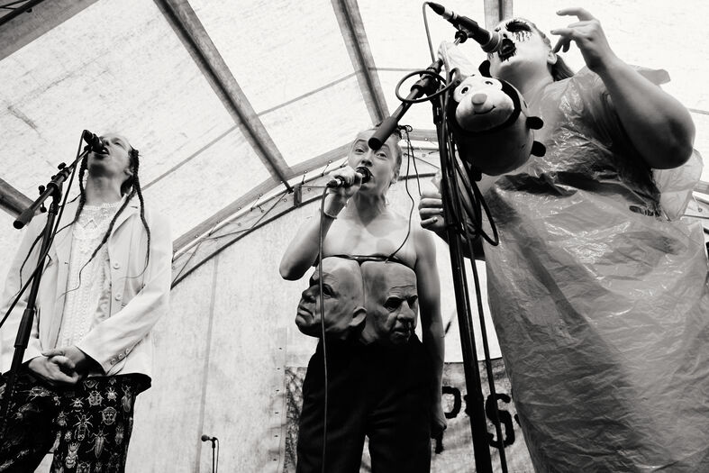 Drie performers met microfoons op een podium, gekleed in opvallende outfits, onder een doorzichtig tentzeil.