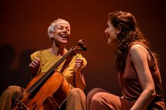 Twee vrouwen lachen en zitten samen op een podium, één met een cello en de ander in een bruine outfit, tegen een warme achtergrond.