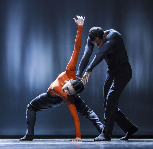 Dansers in zwarte en oranje kostuums voeren een dynamische choreografie uit op een podium met een donkere achtergrond.