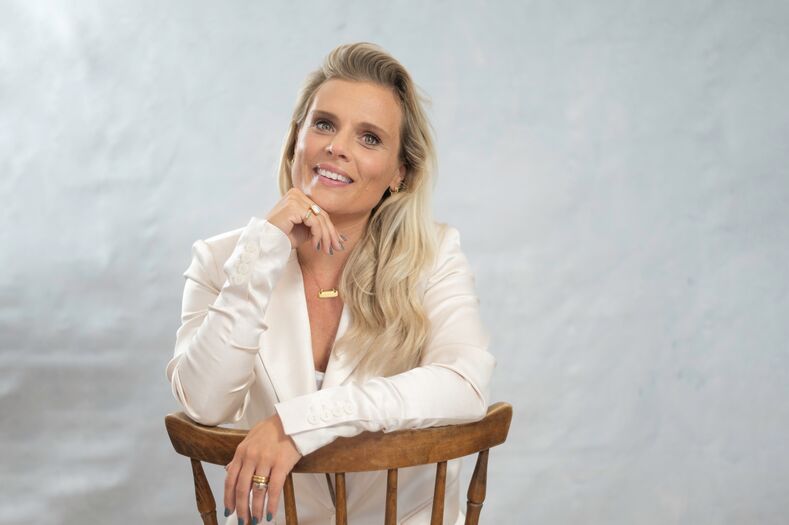 Vrouw met lange blonde haren in witte blouse, zittend op een houten stoel met een glimlach.