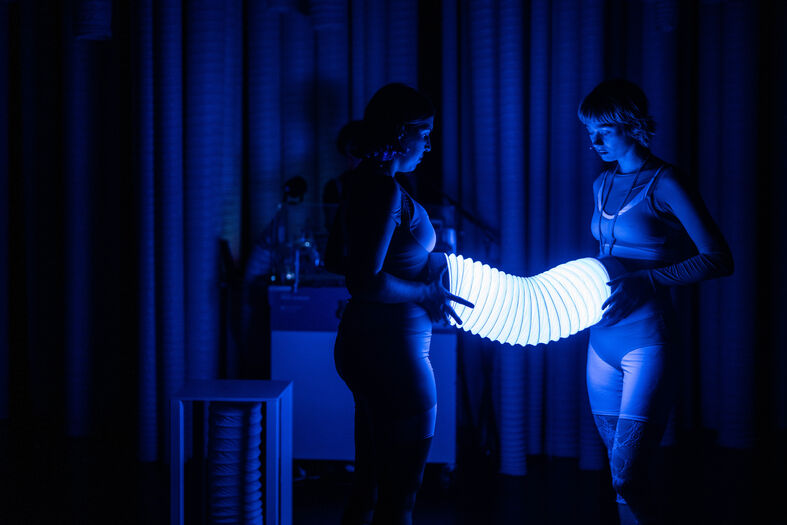 Twee vrouwen in lingerie houden een lichtgevend object vast in een verduisterde ruimte met blauwe verlichting.