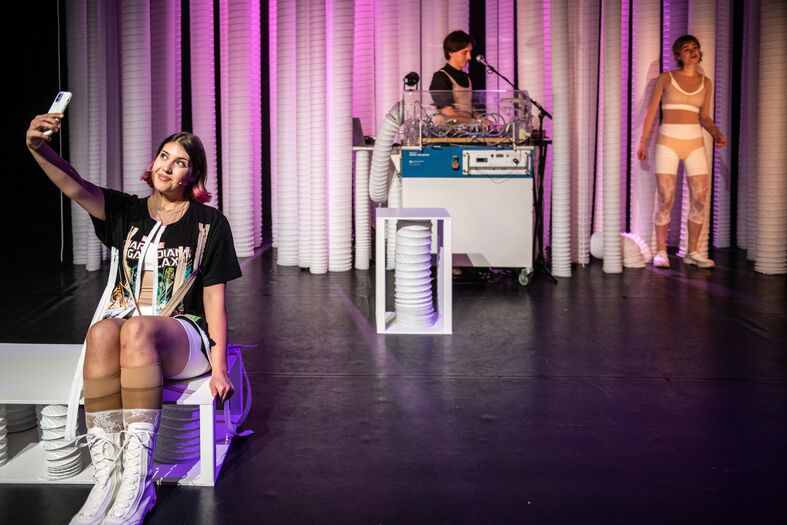 Drie performers op een podium met een muzikale setup; een maakt een selfie terwijl de anderen interageren.