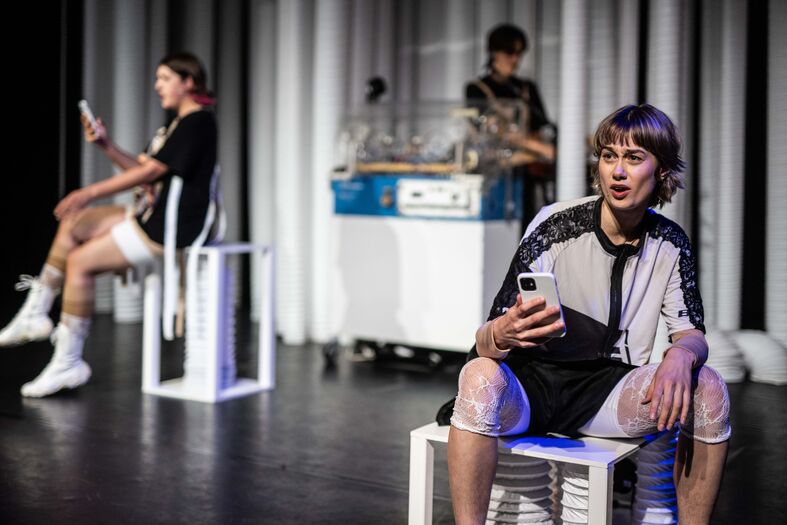Drie jonge mannen in sportkleding zitten op witte stoelen op een podium, met mobiel in hand en een apparaat op de achtergrond.