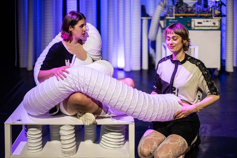 Twee performers op een podium, bezig met een creatieve act met een lange, witte flexibele tube en een tafel.