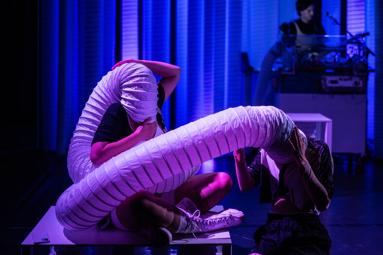 Twee performers op een podium vormen een abstracte beweging met een lange, buigzame constructie onder gedimd licht.