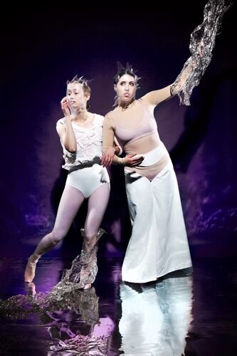 Twee performers in artistieke outfits poseren tegen een paarse achtergrond, met dramatische poses en expressieve gezichten.