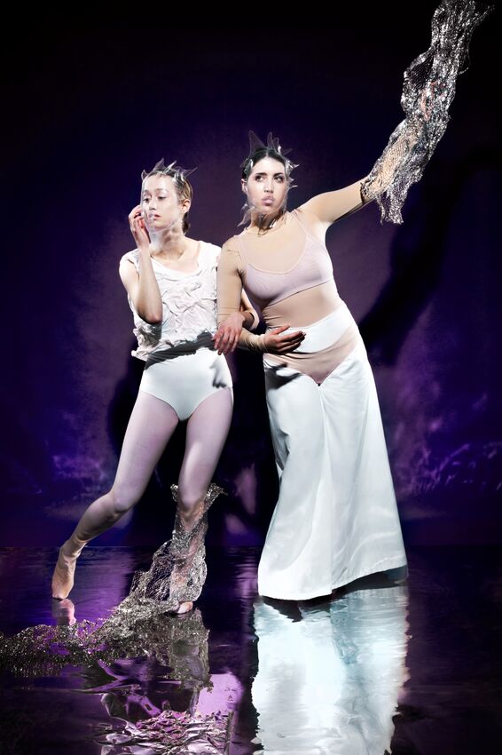 Twee performers in artistieke outfits poseren tegen een paarse achtergrond, met dramatische poses en expressieve gezichten.