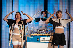 Twee vrouwelijke performers dansen op het podium met een muzikant achter een DJ-set, kleurrijke verlichting op de achtergrond.