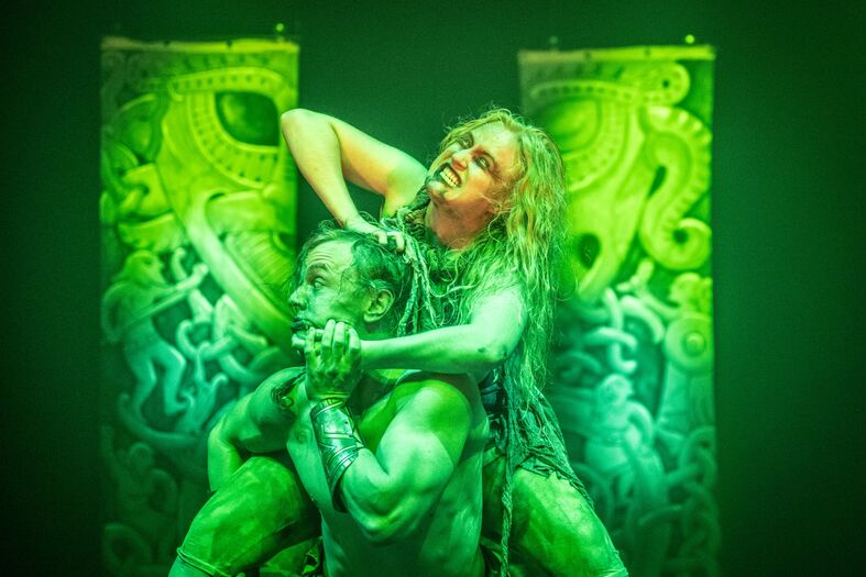 Twee worstelaars met groene lichaamsverf voeren een act uit, omringd door groene achtergronddoeken met octopusmotieven.