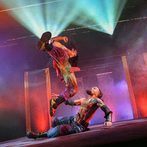 Twee worstelaars voeren een spectaculaire stunt uit op het podium, verlicht door kleurrijke theatrale lichten.