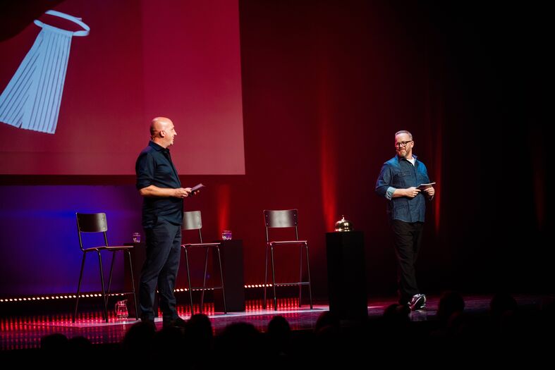 Twee sprekers staan op een podium met een projector op de achtergrond, tijdens een presentatie of evenement.