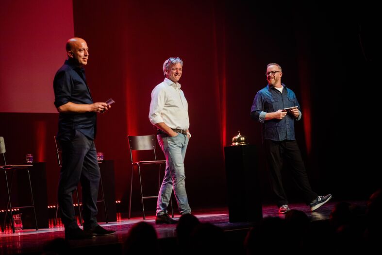Drie sprekers staan op een podium met een rode achtergrond tijdens een evenement, terwijl ze naar het publiek kijken.