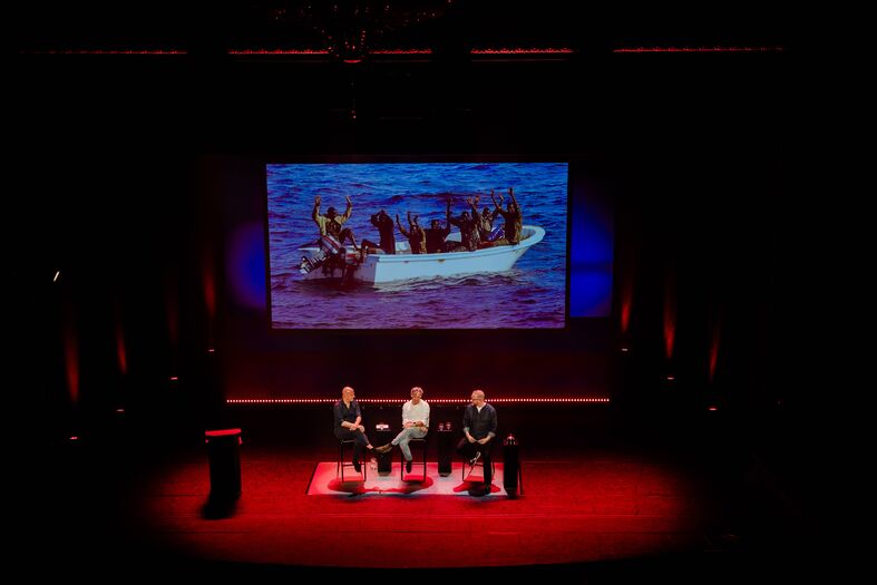 Paneldiscussie op een podium met drie sprekers, terwijl een video van een boot met mensen op de achtergrond wordt getoond.