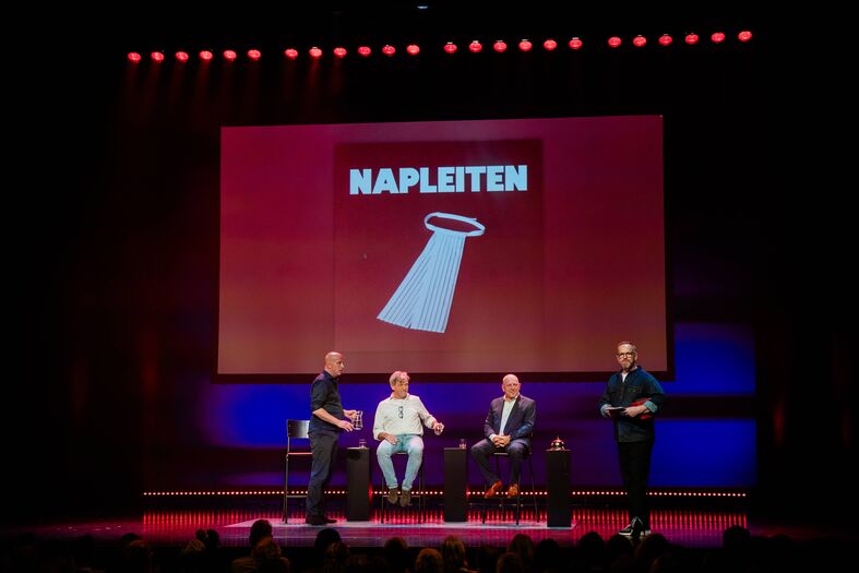 Drie sprekers op een podium tijdens een evenement, met een groot scherm op de achtergrond waarop "NAPLETTEN" staat.