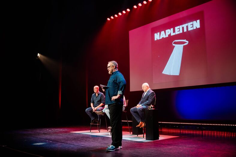 Twee sprekers op een podium met een presentatie achter hen, getiteld "NAPLETEN". Verlichting in warme kleuren.
