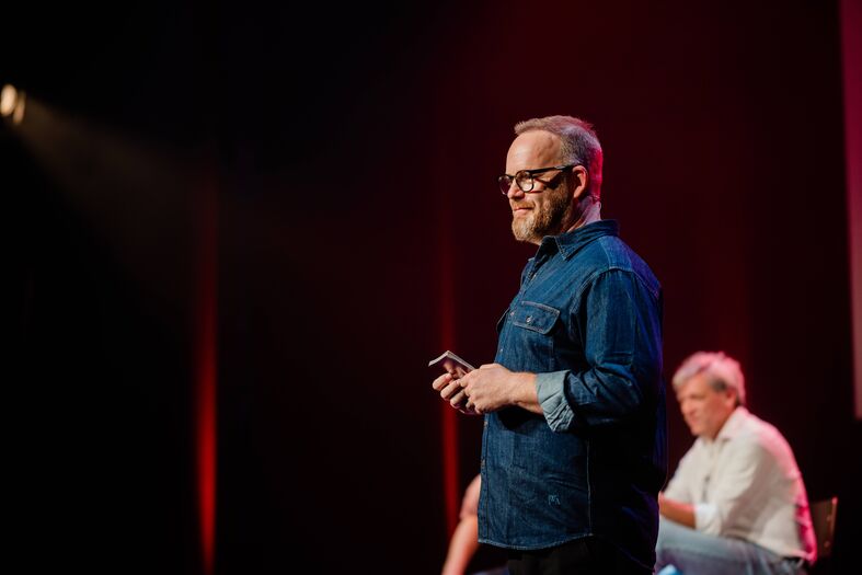Man met een bril en een blauwe blouse staat op het podium tijdens een presentatie, met een andere spreker op de achtergrond.