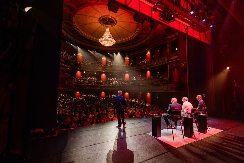 Een podium met sprekers voor een publiek in een theater met rijke decoraties en een kroonluchter.