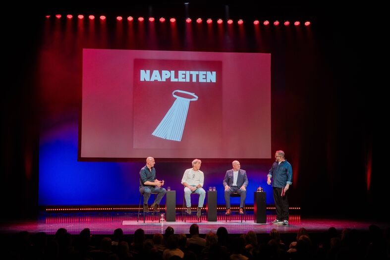 Drie sprekers op een podium voor een scherm met de tekst "NAPLETEN" en een afbeelding van een zoutvaatje.