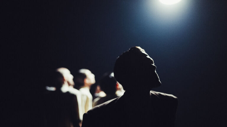 Meerdere silhouetten staan in donker podiumlicht terwijl één persoon omhoog kijkt naar felle spot.