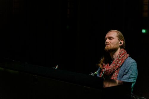 Man met een sjaal speelt piano in een donkere ruimte, met een serene uitstraling.
