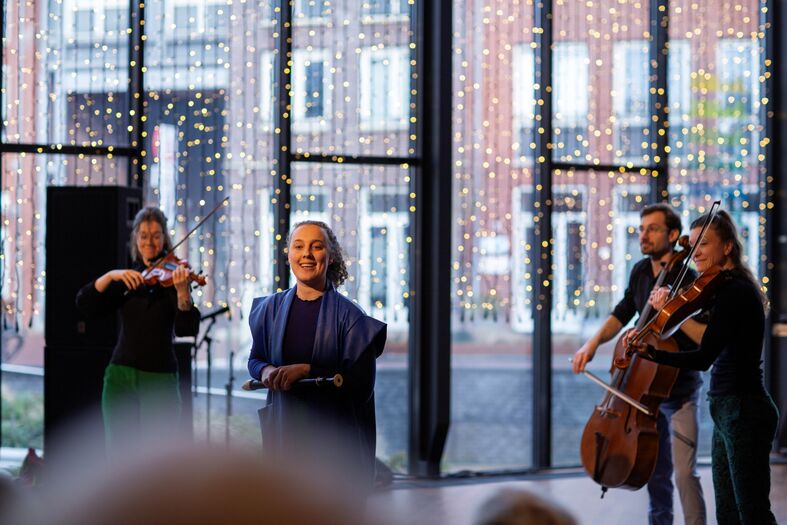 Een muzikale bijeenkomst met een spreker en drie musici, waaronder een violist, cellist en een zangeres, in een sfeervolle ruimte.